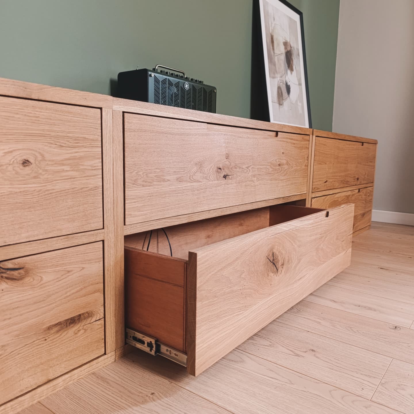 Długa, niska komoda z jasnego drewna dębowego z otwartą szufladą, ukazującą ciemniejsze wnętrze. Na komodzie głośnik i grafika w czarnej ramie.