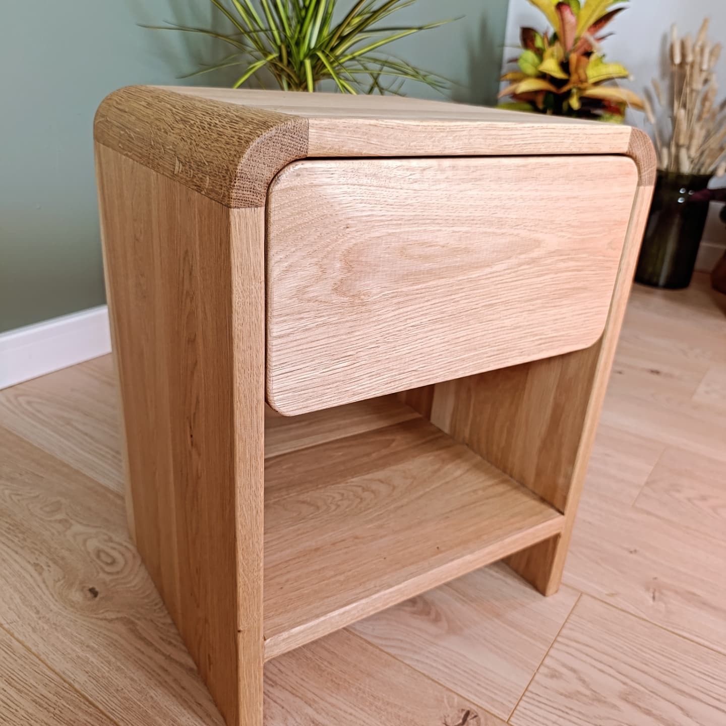 Drewniana szafka nocna z szufladą i półką, zaokrąglone krawędzie, naturalne wykończenie drewna dębowego, na tle roślin w doniczkach.