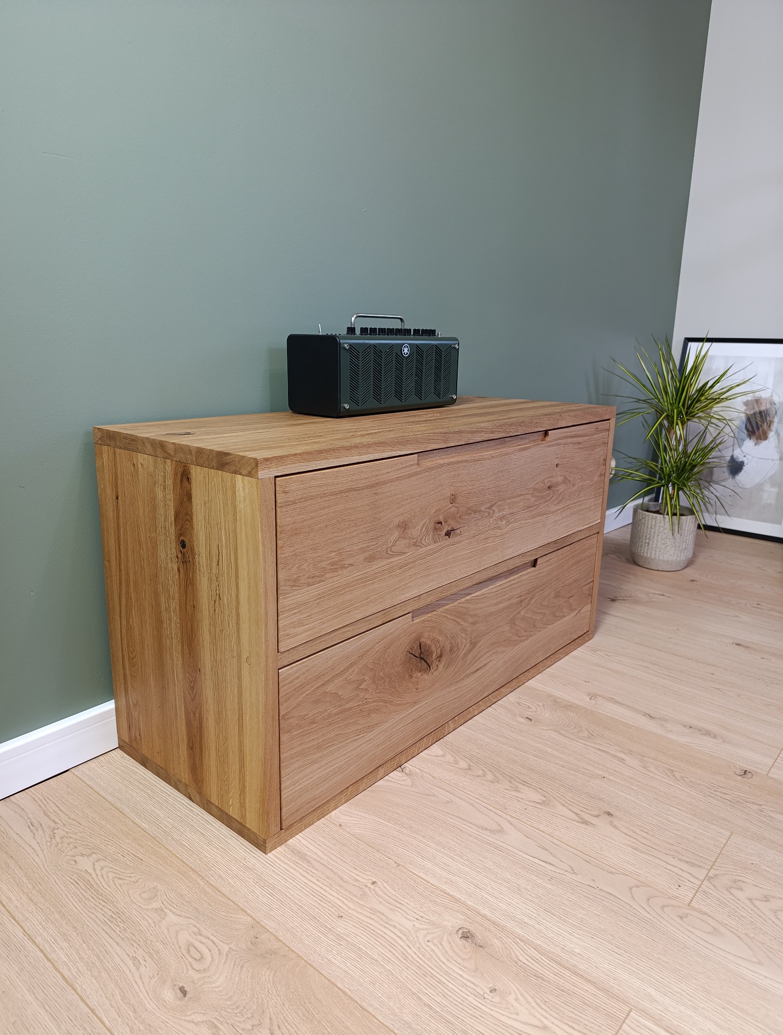 Dębowa komoda z dwoma szufladami, na blacie głośnik Yamaha. Widoczne słoje drewna i detale wykończenia. W tle ściana w kolorze oliwkowym i roślina doniczkowa.