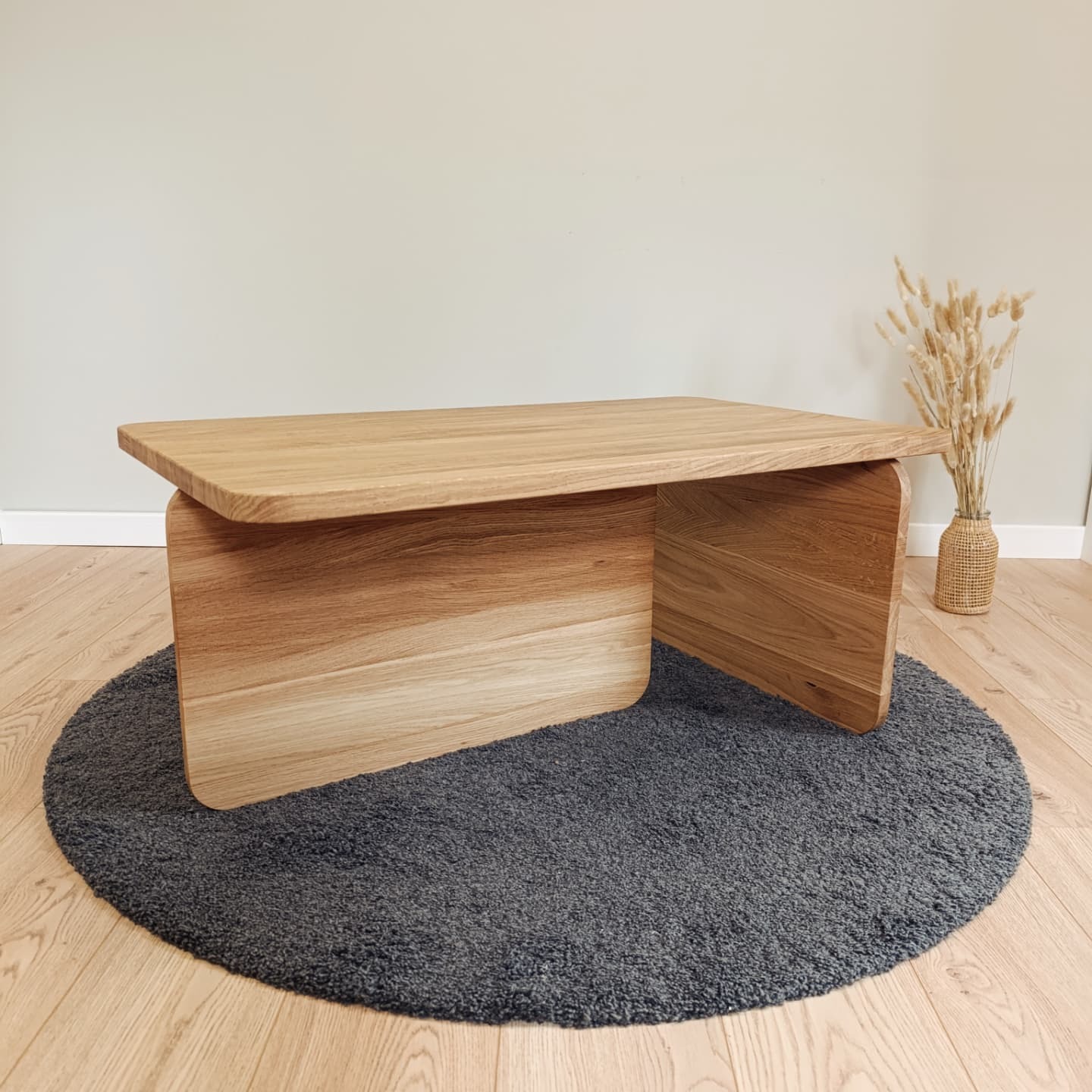 Stolik kawowy z jasnego drewna o minimalistycznym designie, stojący na okrągłym, szarym dywanie. W tle wazon z suszonymi trawami.