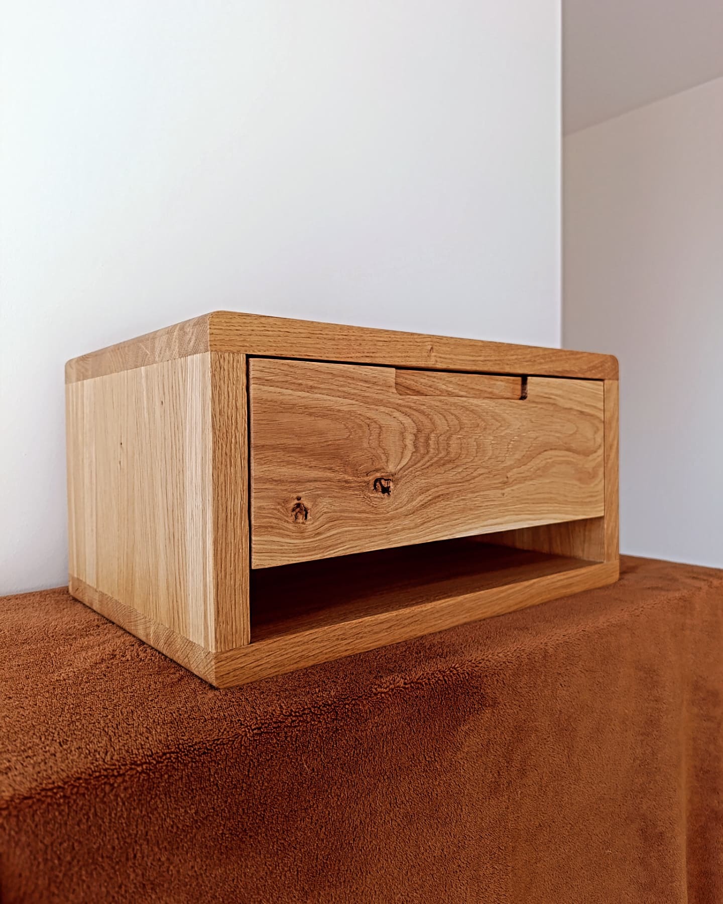 Drewniana szafka nocna z szufladą, wykonana z jasnego drewna dębowego, z widocznymi słojami i naturalnymi sękami, na tle białej ściany i brązowego materiału.