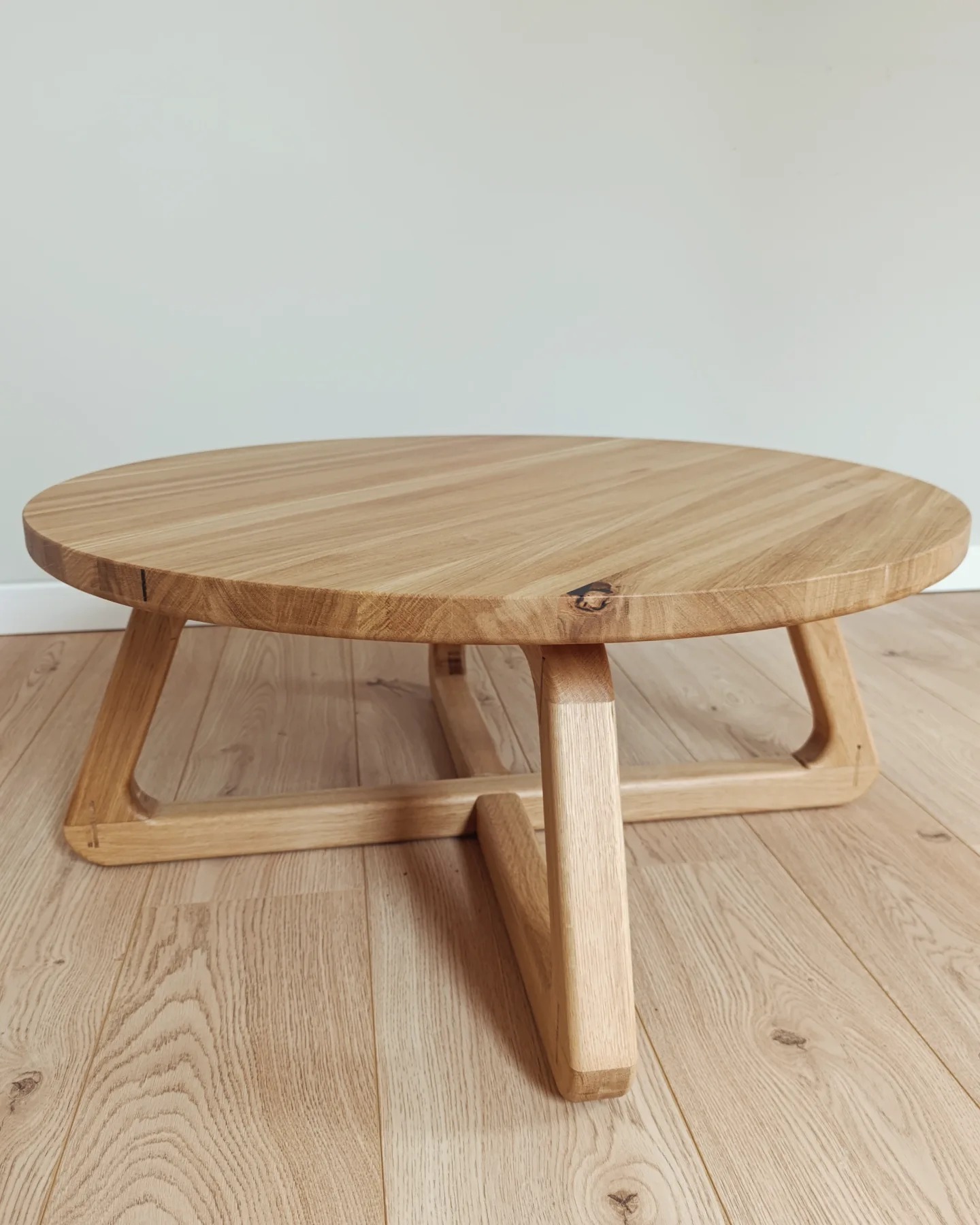 Okrągły stolik kawowy z jasnego drewna z widocznym usłojeniem, na tle podłogi z paneli i jasnej ściany. Niskie nóżki w geometrycznym stylu.