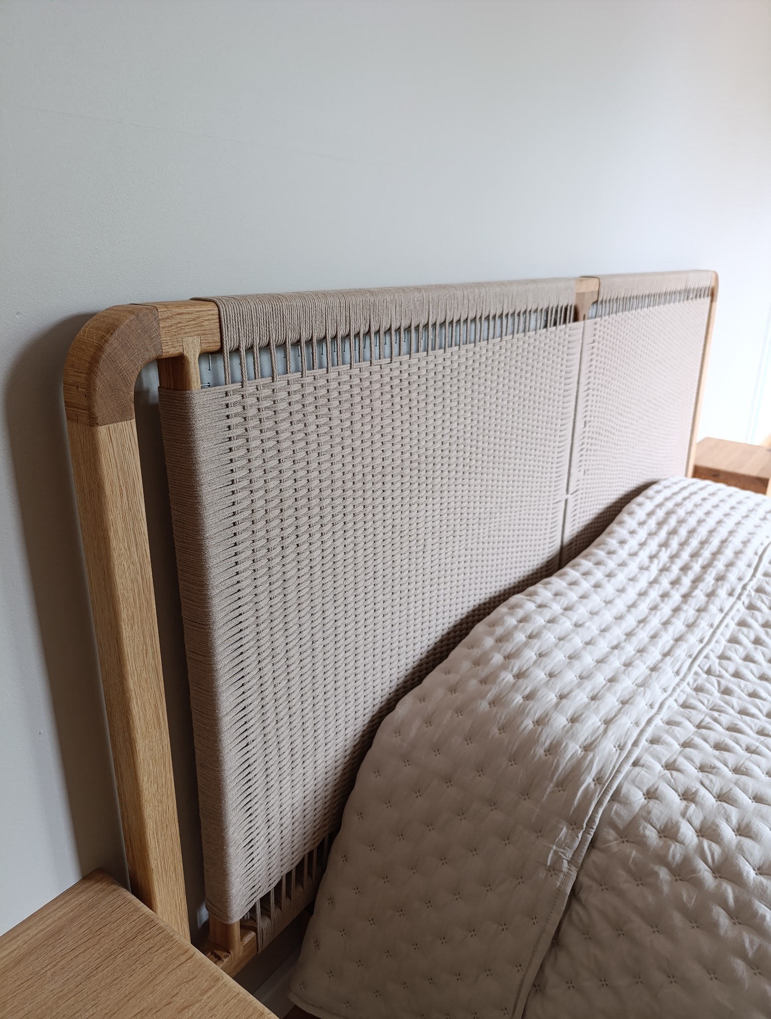 Ręcznie pleciony zagłówek łóżka z jasnego drewna i beżowego sznurka, widoczny fragment szafki nocnej i kołdry w neutralnych barwach, minimalistyczny design.