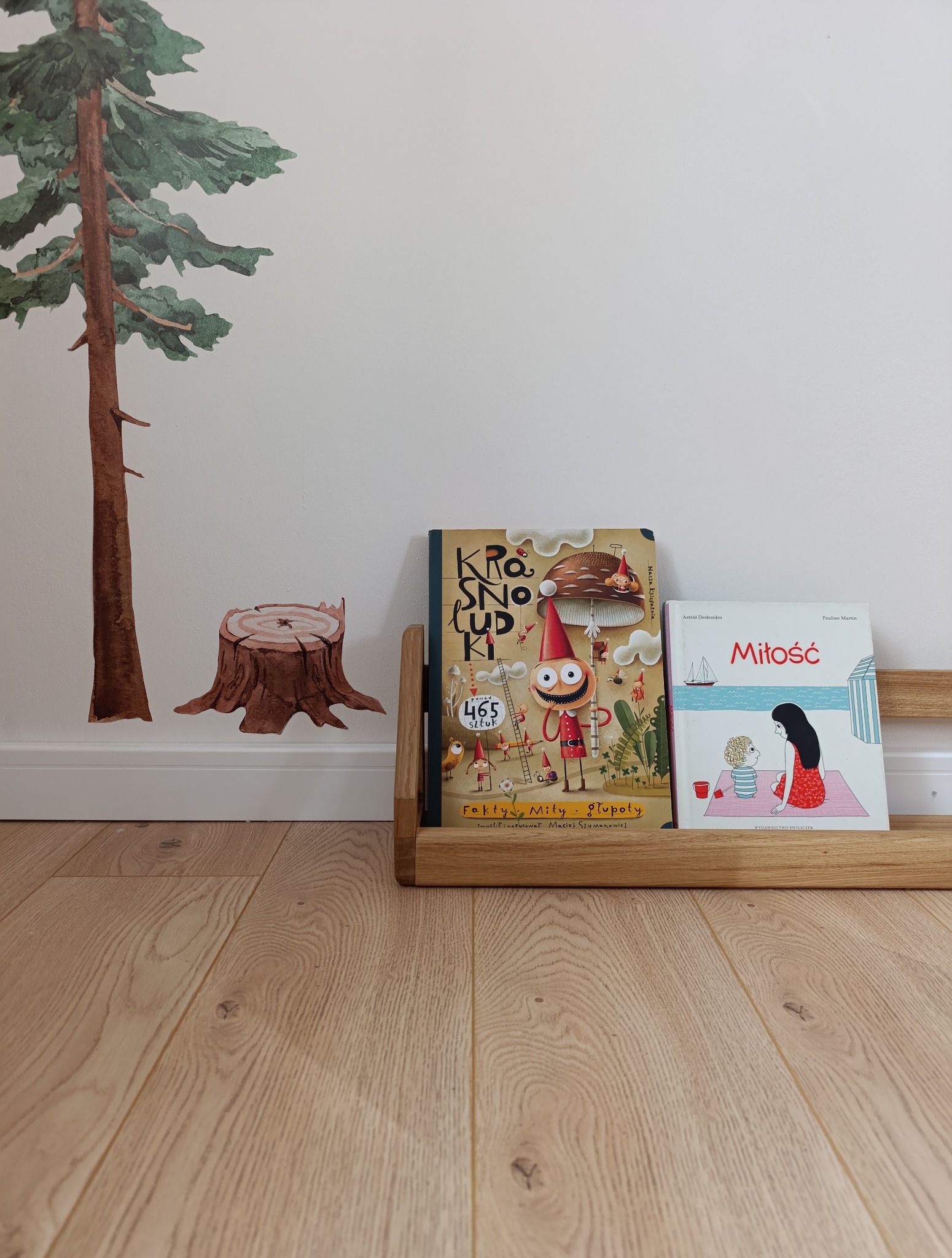 Drewniana półka na książki dziecięce, z widoczną okleiną dębową i dwiema książkami. Na ścianie naklejki drzewa i pnia. Podłoga z desek.