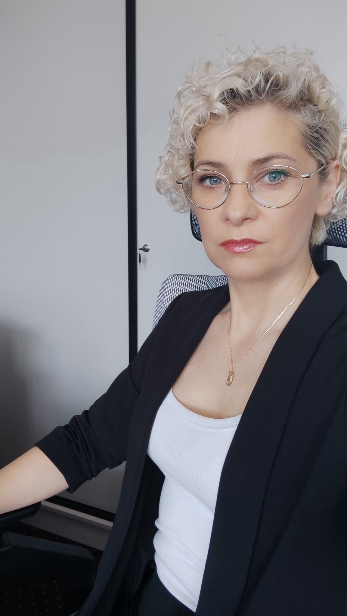 Portret kobiety w okularach, z blond kręconymi włosami, w czarnej marynarce i białym topie, siedzącej przy biurku w biurze. Spokojny wyraz twarzy, złoty łańcuszek.