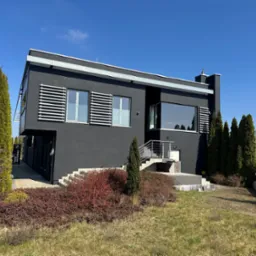 Nowoczesny budynek z ciemną elewacją i białymi żaluzjami okiennymi, otoczony zielenią i błękitnym niebem. Widoczne schody wejściowe i zadbany trawnik.