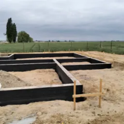 Zabezpieczone fundamenty domu z czarną izolacją, widoczne drewniane podpory i przygotowany teren pod budowę na tle zielonego pola i pochmurnego nieba.