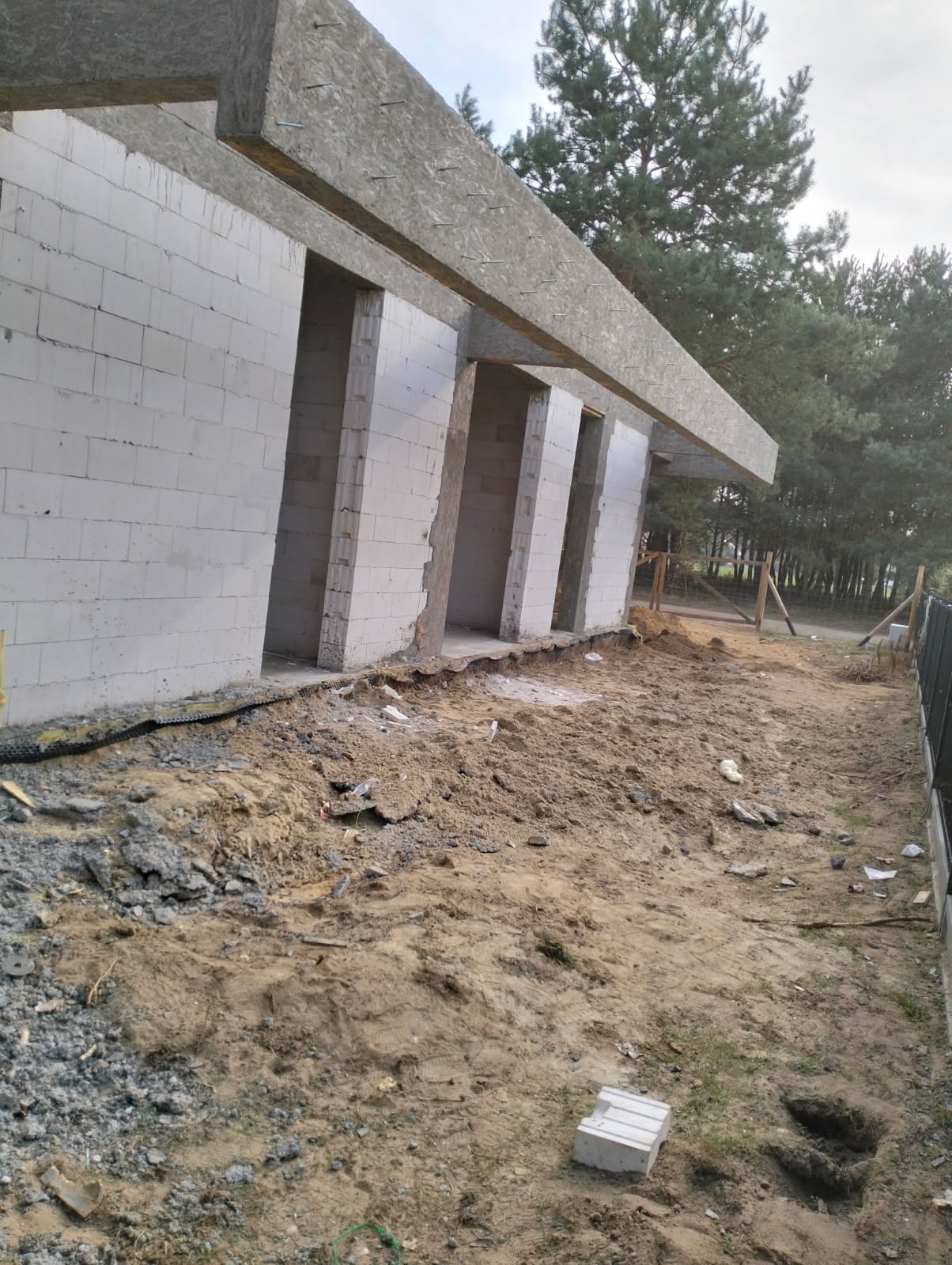 Wznoszenie ścian z betonu komórkowego Ytong. Widoczne otwory okienne i drzwiowe. Częściowo zadaszona konstrukcja. Na pierwszym planie ziemia i pojedynczy bloczek.