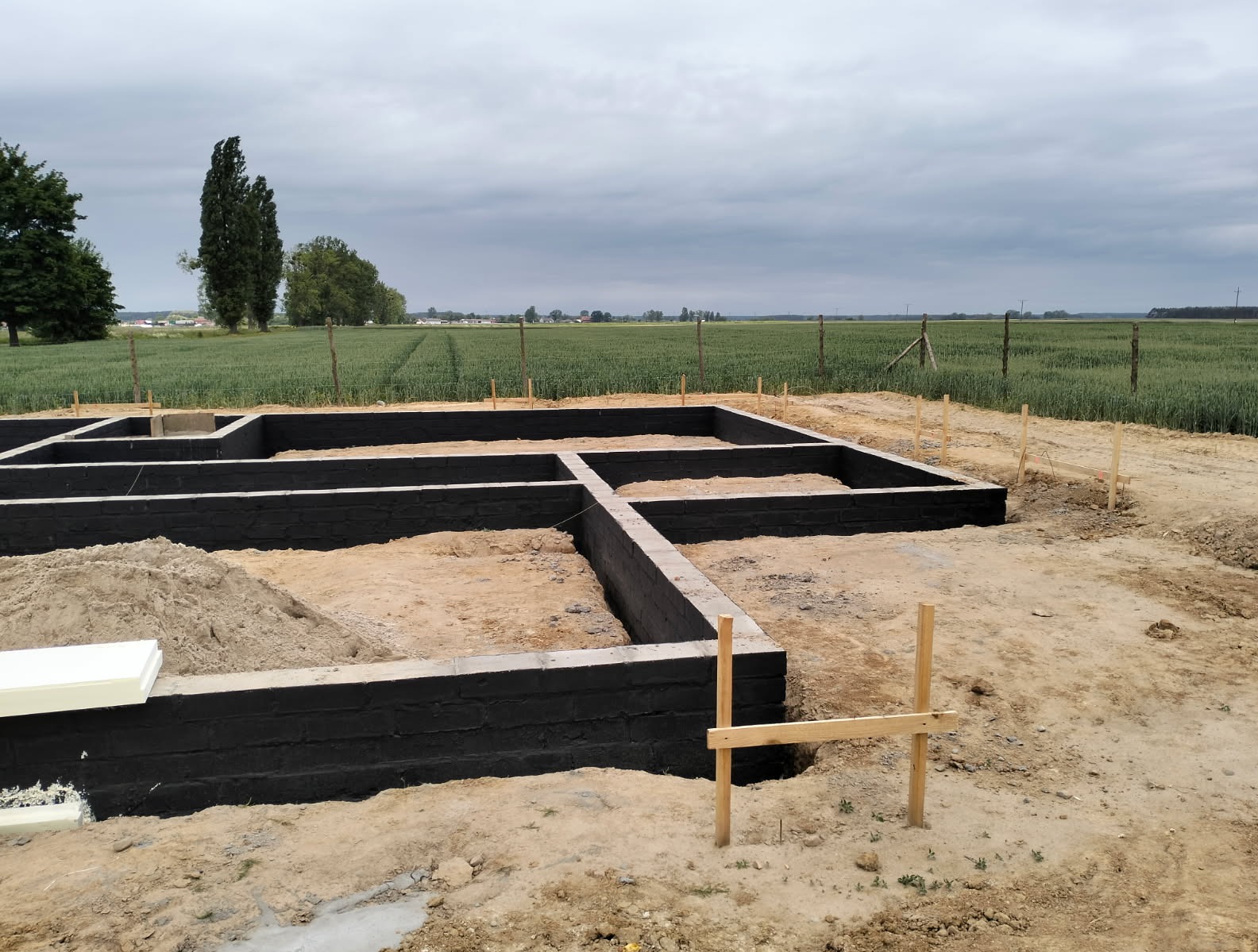Zabezpieczone fundamenty domu z czarną izolacją, widoczne drewniane podpory i przygotowany teren pod budowę na tle zielonego pola i pochmurnego nieba.