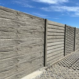 P.H.U. M-R-B Therm inż. Mateusz BUDZIŃSKI - Długi, szary płot betonowy z różnymi wzorami paneli (imitacja drewna, poziome linie i fale) na tle błękitnego nieba. Widoczna kostka ażurowa.