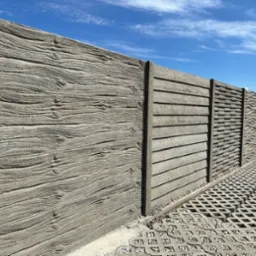 Długi, szary płot betonowy z różnymi wzorami paneli (imitacja drewna, poziome linie i fale) na tle błękitnego nieba. Widoczna kostka ażurowa.