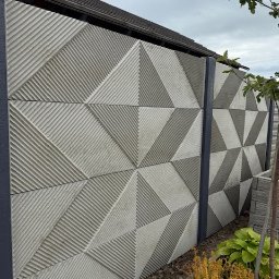 P.H.U. M-R-B Therm inż. Mateusz BUDZIŃSKI - Geometryczny wzór na betonowym ogrodzeniu panelowym z szarymi słupkami, zieleń w donicach u podstawy. Nowoczesny design.