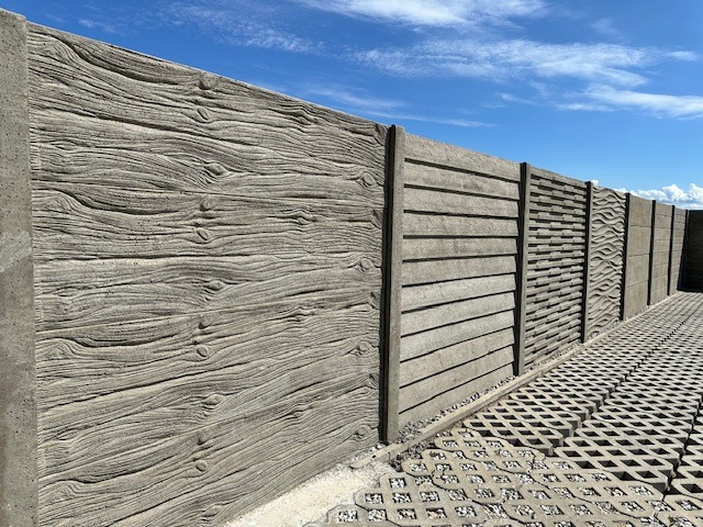 Długi, szary płot betonowy z różnymi wzorami paneli (imitacja drewna, poziome linie i fale) na tle błękitnego nieba. Widoczna kostka ażurowa.