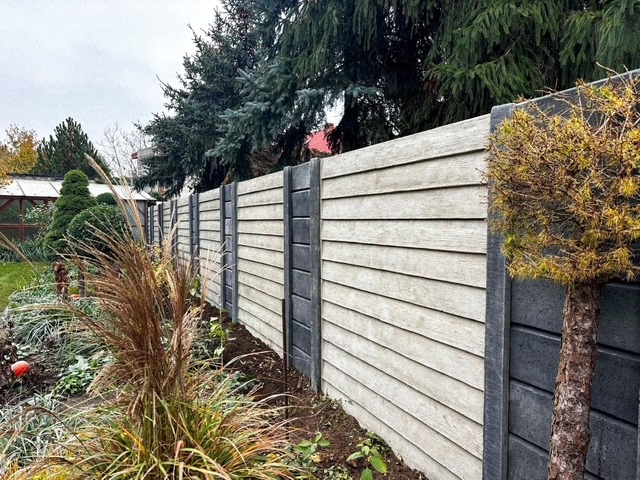 Nowoczesne ogrodzenie z paneli imitujących drewno, z szarymi słupkami, otoczone zielenią ogrodu, w tle widoczne drzewa iglaste.