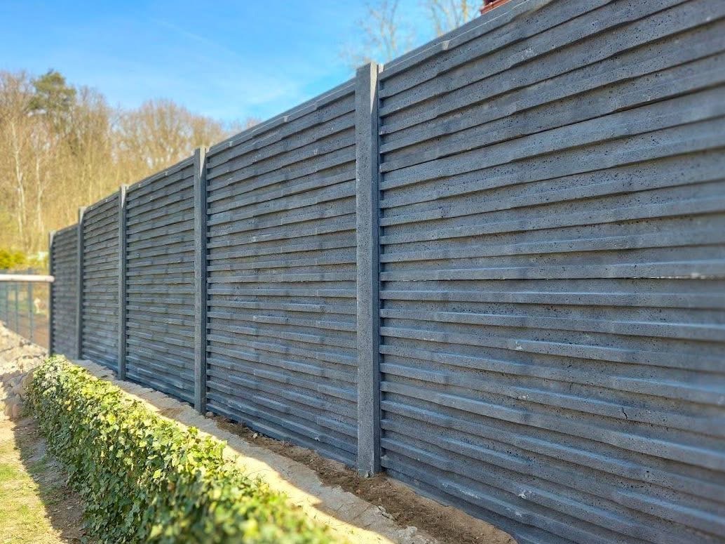 Nowoczesne, szare ogrodzenie betonowe z poziomymi panelami, oddzielające posesję od zieleni. Widoczny fragment trawnika i krzewów. Słoneczny dzień.