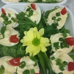 Dekoracyjnie podane jajka faszerowane z majonezem, pomidorem i szczypiorkiem, na zielonej sałacie, z żółtym kwiatem pośrodku, w białym naczyniu.