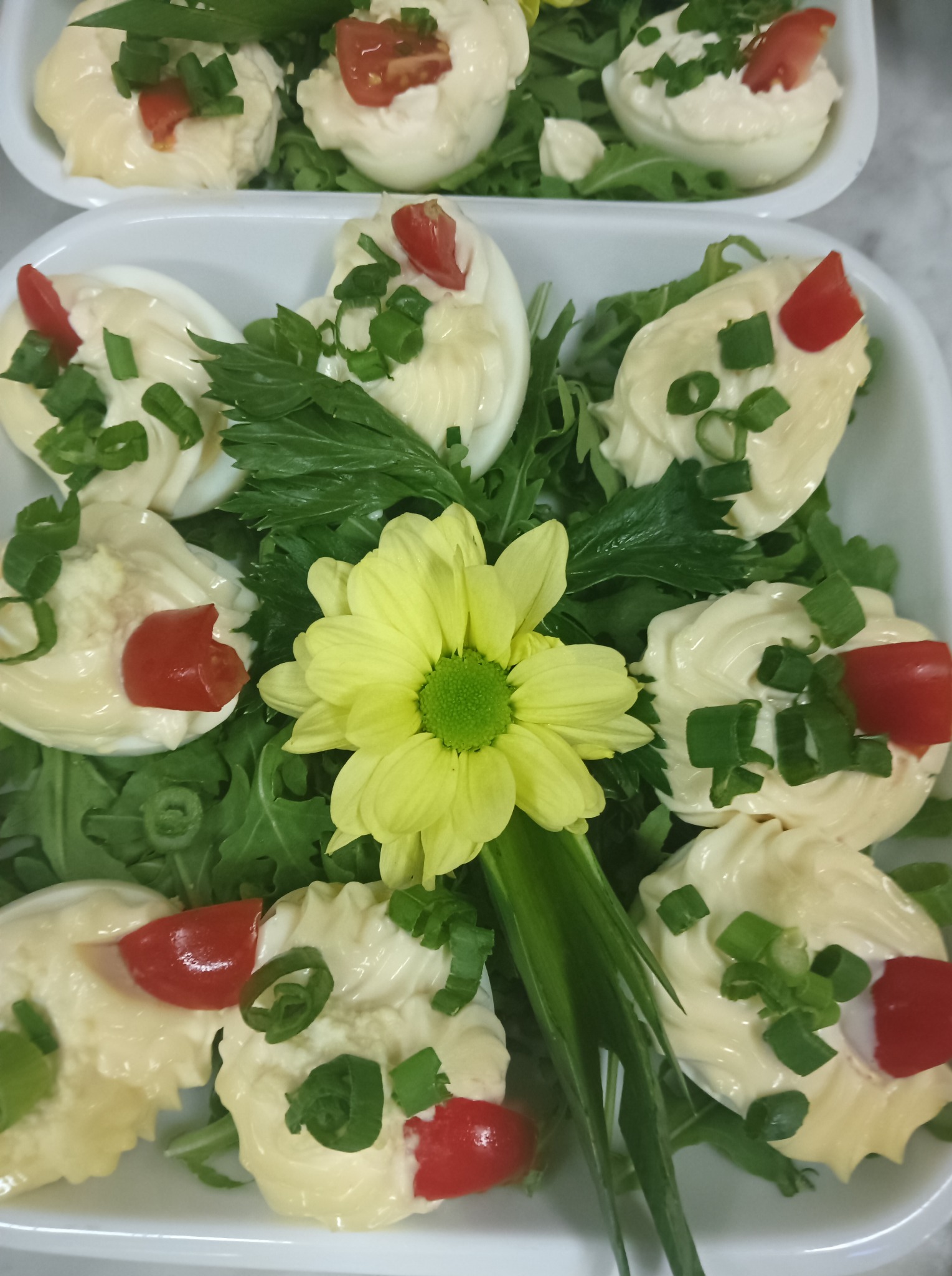 Dekoracyjnie podane jajka faszerowane z majonezem, pomidorem i szczypiorkiem, na zielonej sałacie, z żółtym kwiatem pośrodku, w białym naczyniu.