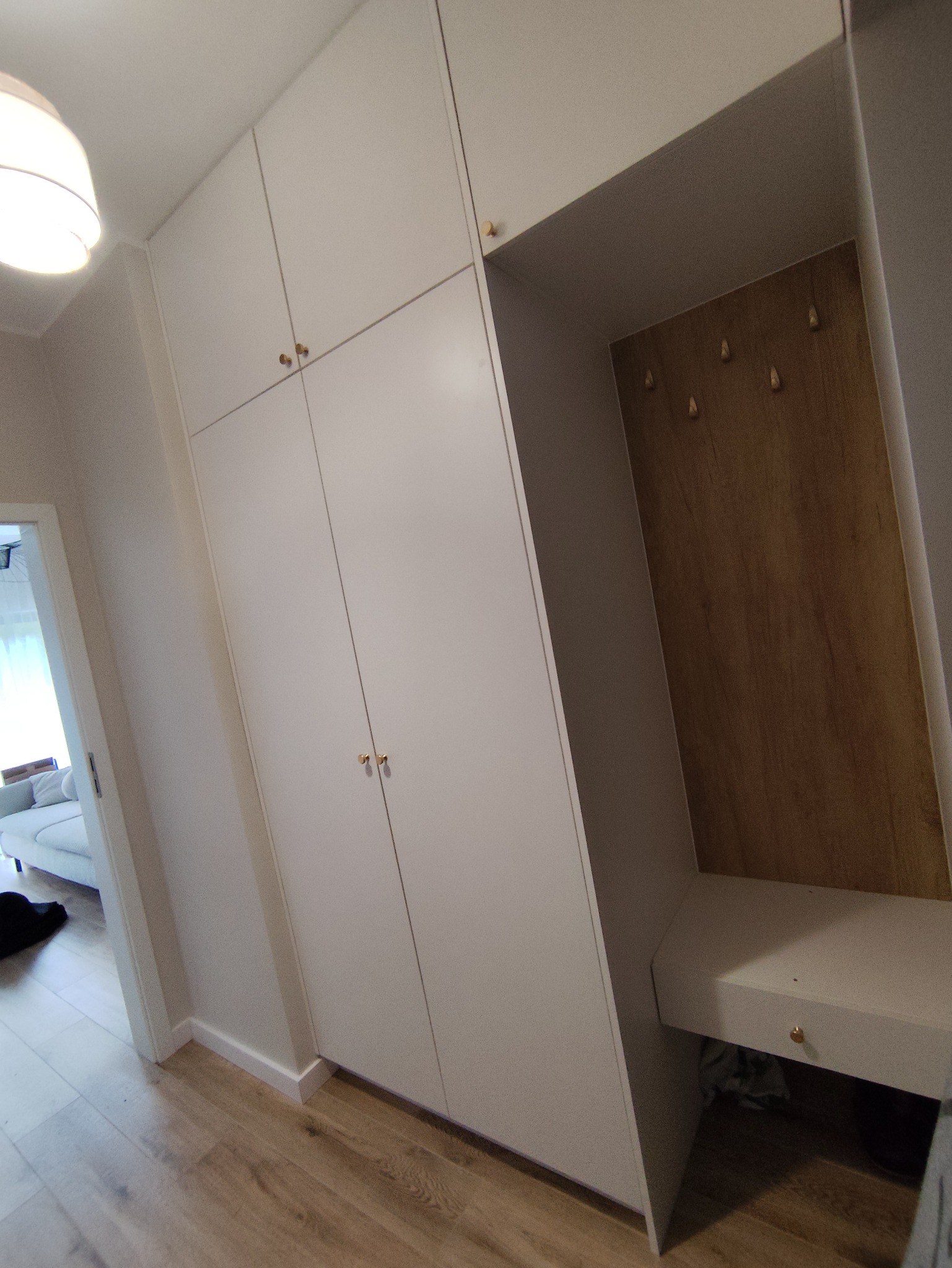 Biała szafa wnękowa z drewnianym panelem i siedziskiem w przedpokoju. Złote uchwyty dodają elegancji. Widoczna podłoga z paneli.