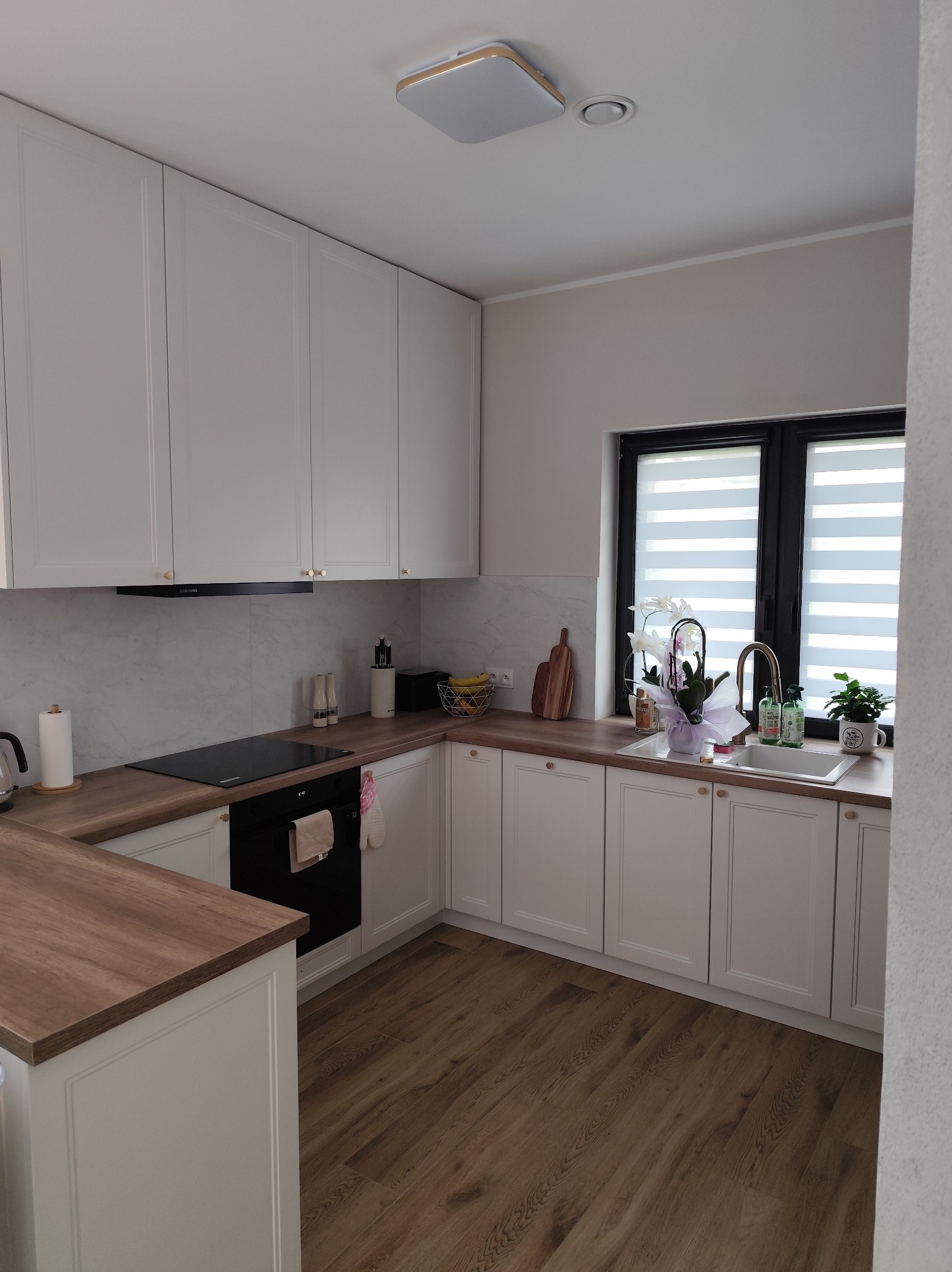 Jasna kuchnia w stylu klasycznym z drewnianym blatem, białymi szafkami i czarnymi akcentami, jak okno i płyta grzewcza. Uwagę przyciągają złote uchwyty.