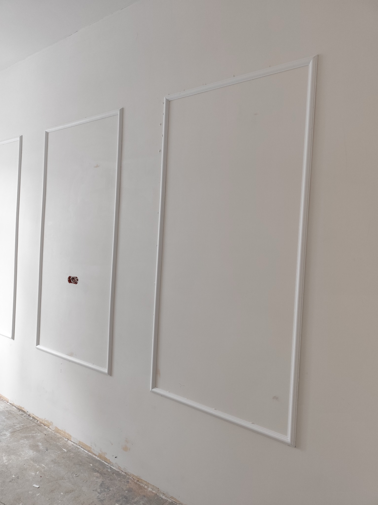 Ściana z dekoracyjnymi, białymi ramkami. Widoczne ślady szpachlowania i gniazdko elektryczne w jednej z ramek. Surowa podłoga.