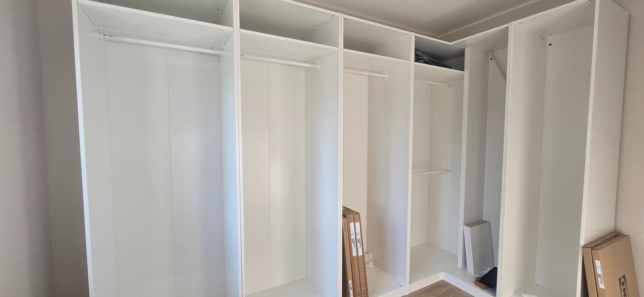 Biała, modułowa szafa wnękowa z drążkami i półkami, jeszcze bez drzwi, w trakcie montażu. Widoczne elementy montażowe i kartony. Ujęcie z dołu, szeroki kąt.