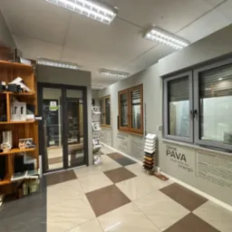 Ekspozycja okien i rolet w salonie: okna drewniane, aluminiowe i PCV, rolety zewnętrzne szare, próbki kolorów. Wnętrze z szachownicową podłogą.