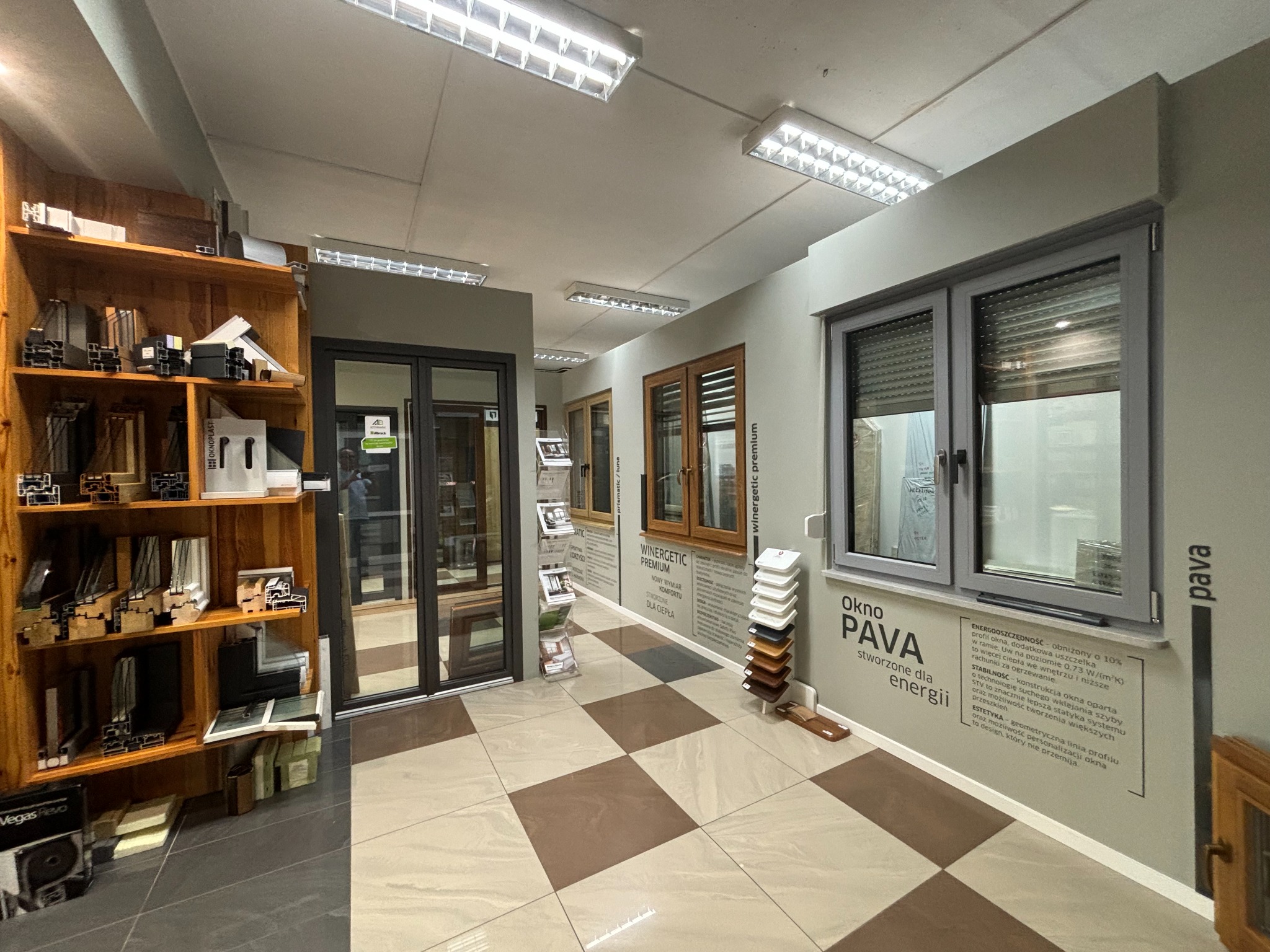 Ekspozycja okien i rolet w salonie: okna drewniane, aluminiowe i PCV, rolety zewnętrzne szare, próbki kolorów. Wnętrze z szachownicową podłogą.