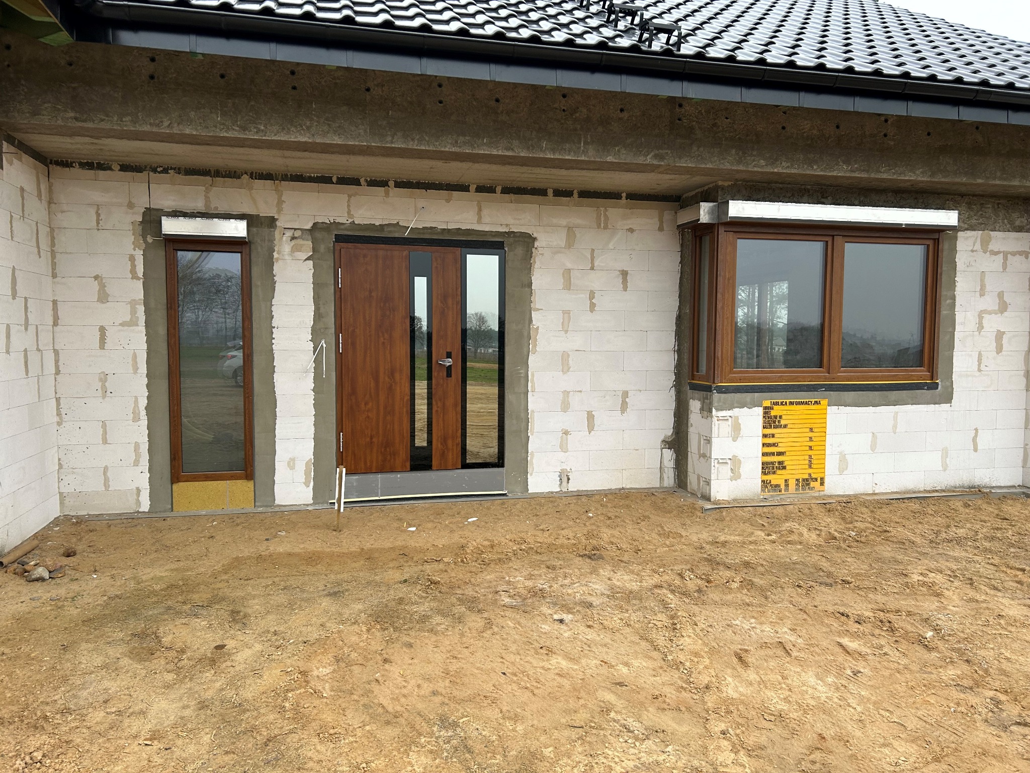 Nowy dom w trakcie budowy: zamontowane brązowe okna i drzwi wejściowe z czarnymi ramami w surowym murze z bloczków. Widoczna tablica informacyjna budowy.