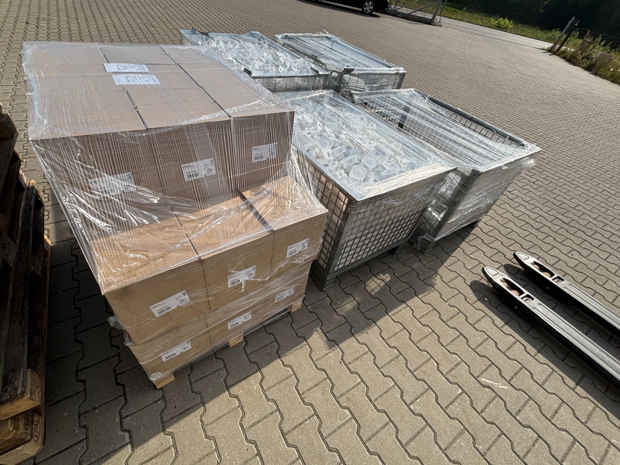 Kartonowe pudła owinięte folią i metalowe kosze z zawartością, ustawione na paletach, gotowe do transportu. Widoczny wózek paletowy i kostka brukowa.