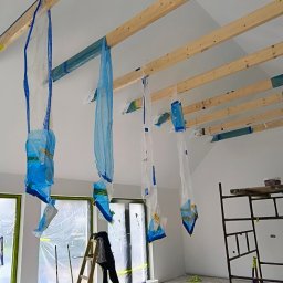 Daniel Lebok - Remont pokoju: zabezpieczone okna folią, worki na suficie, drabina i rusztowanie w pomieszczeniu, przygotowanym do malowania. Widoczne drewniane belki.