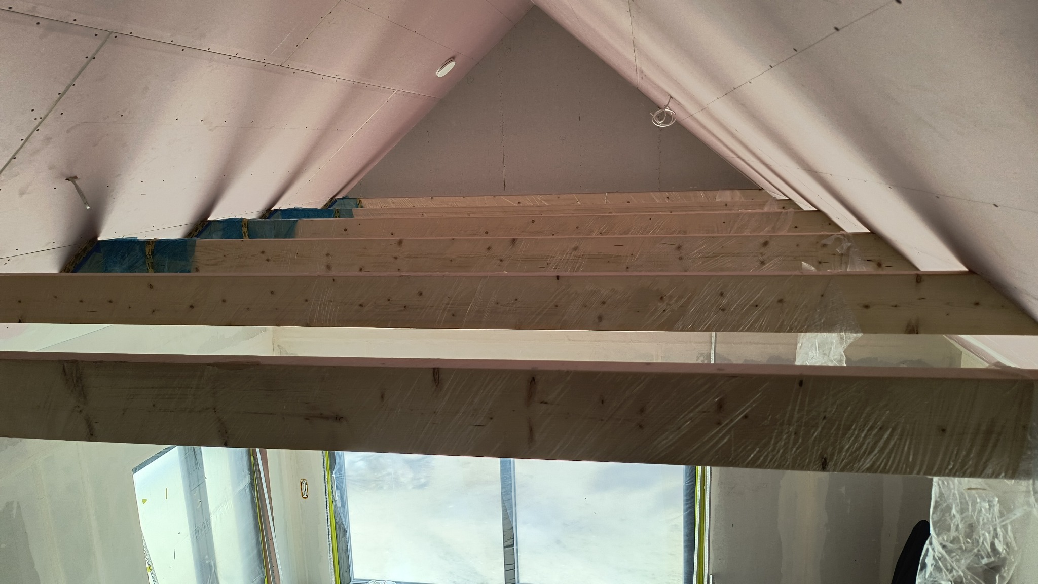Skośny sufit poddasza z płyt karton-gips i drewnianą konstrukcją, zabezpieczoną folią. Widok z dołu na okno dachowe. Prace wykończeniowe wnętrza.