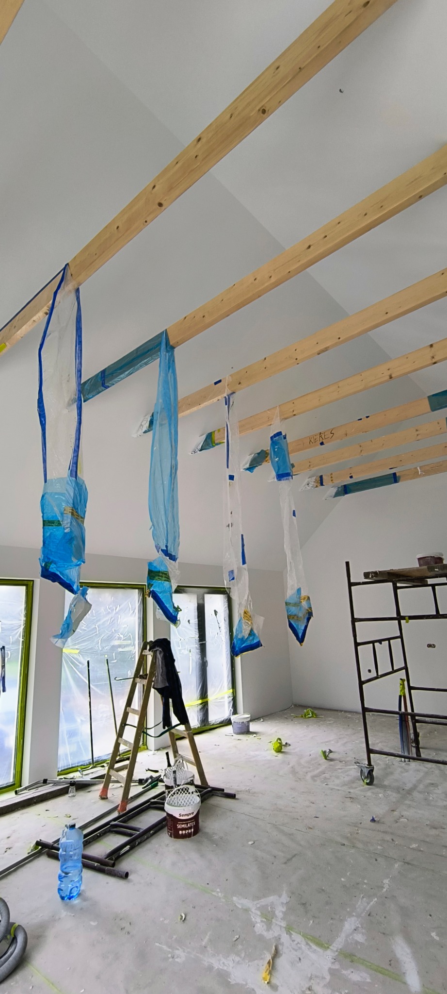 Remont pokoju: zabezpieczone okna folią, worki na suficie, drabina i rusztowanie w pomieszczeniu, przygotowanym do malowania. Widoczne drewniane belki.