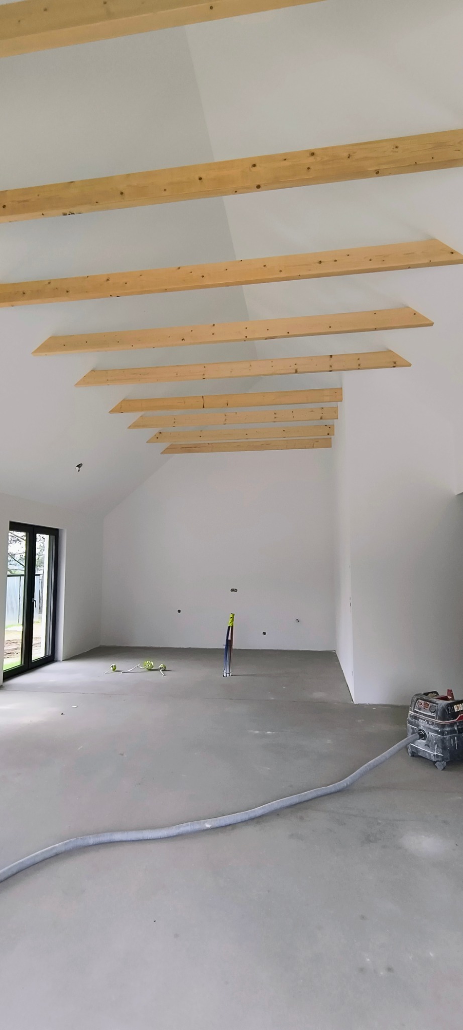 Wnętrze w trakcie prac wykończeniowych: surowa podłoga, białe ściany, drewniane belki na suficie i sprzęt budowlany. Widok na okno i instalacje.