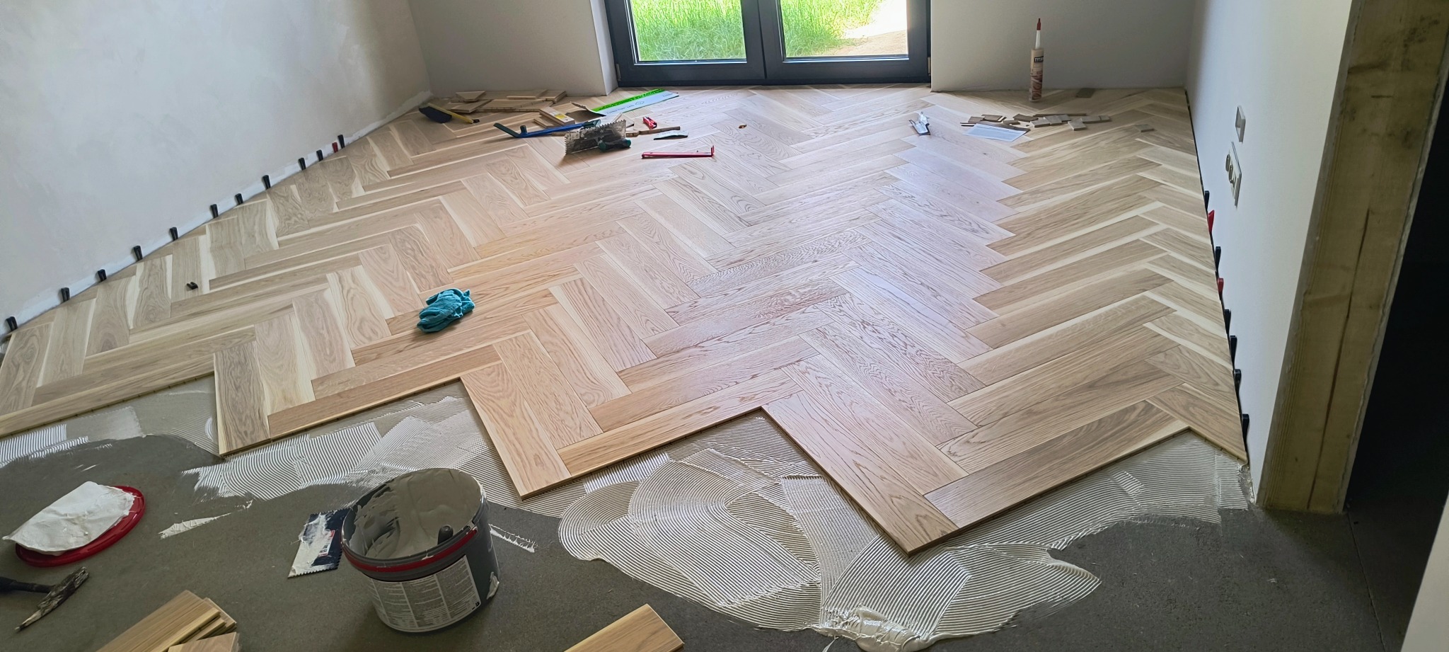 Układanie parkietu w jodełkę: jasne panele ułożone częściowo, widoczne narzędzia, klej i czarne kliny dystansowe wzdłuż ściany. Praca w toku.