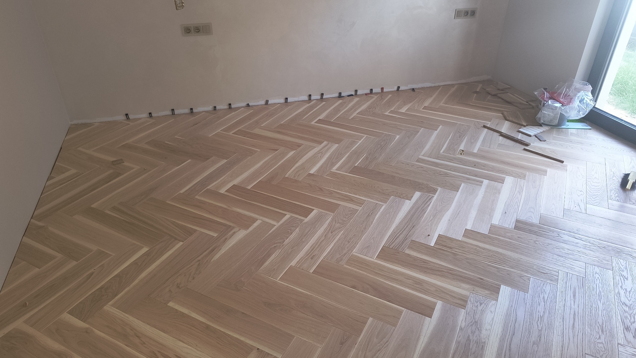 Ułożona parkietowa podłoga w jodełkę w jasnym odcieniu drewna, widoczne listwy dystansowe przy ścianie, okno i materiały montażowe w tle.