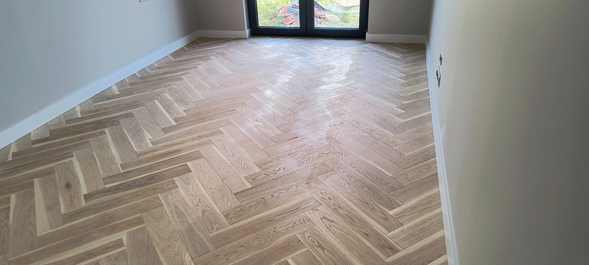 Jasna, drewniana podłoga ułożona w jodełkę francuską w nowym, odświeżonym pokoju z białymi listwami przypodłogowymi i oknem balkonowym.