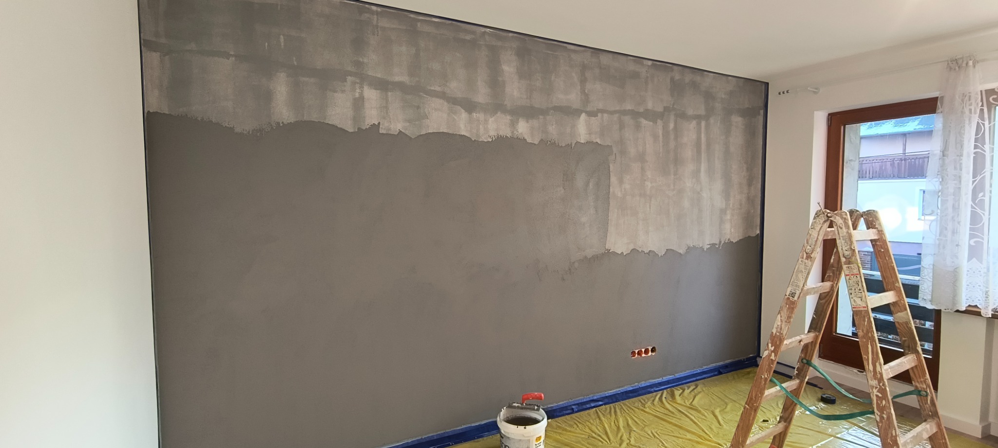 Ściana w trakcie szpachlowania z widoczną drabiną i zabezpieczoną podłogą. Widoczne gniazdka elektryczne i okno z firanką.