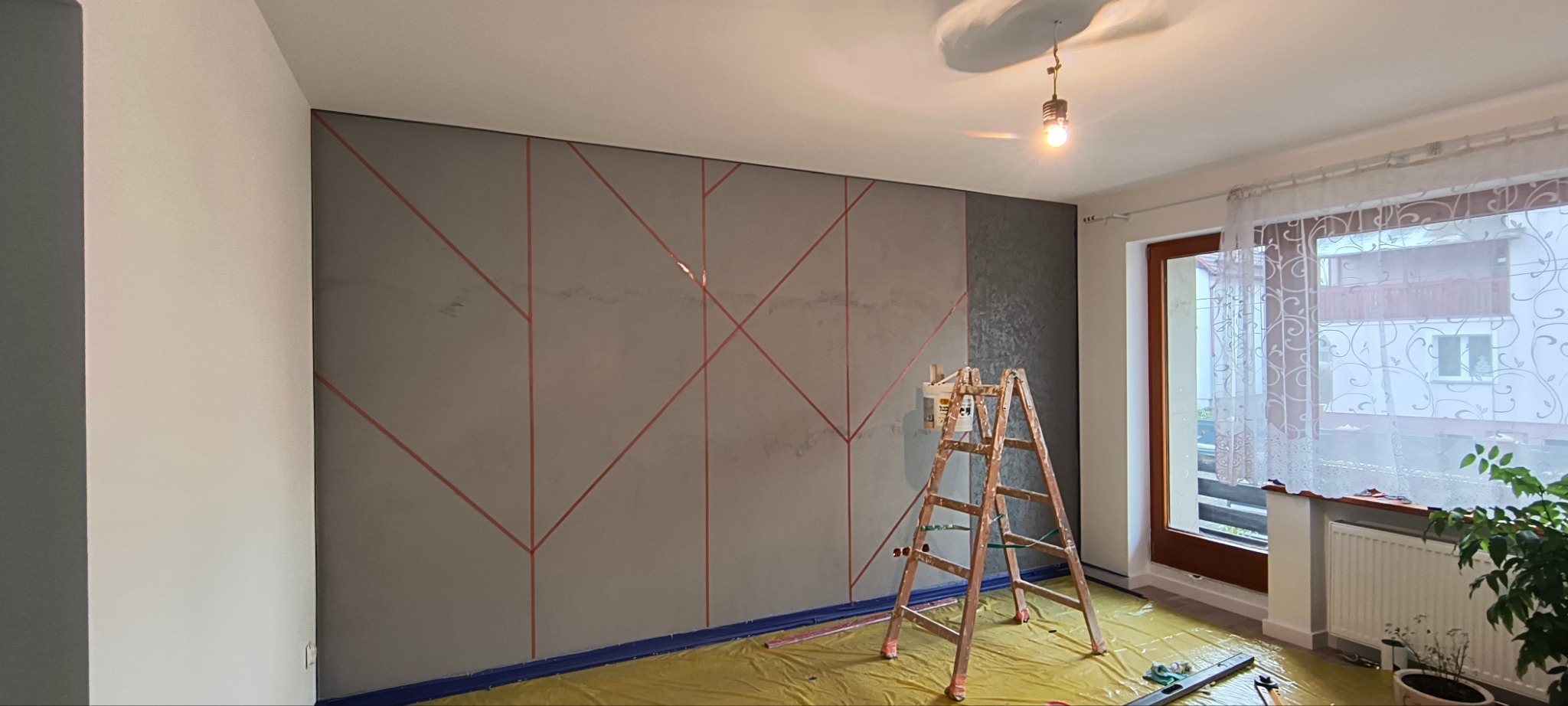 Pokój w trakcie malowania: ściana z geometrycznym wzorem w taśmach, drabina z farbą, podłoga zabezpieczona folią. Widok na okno z firanką.