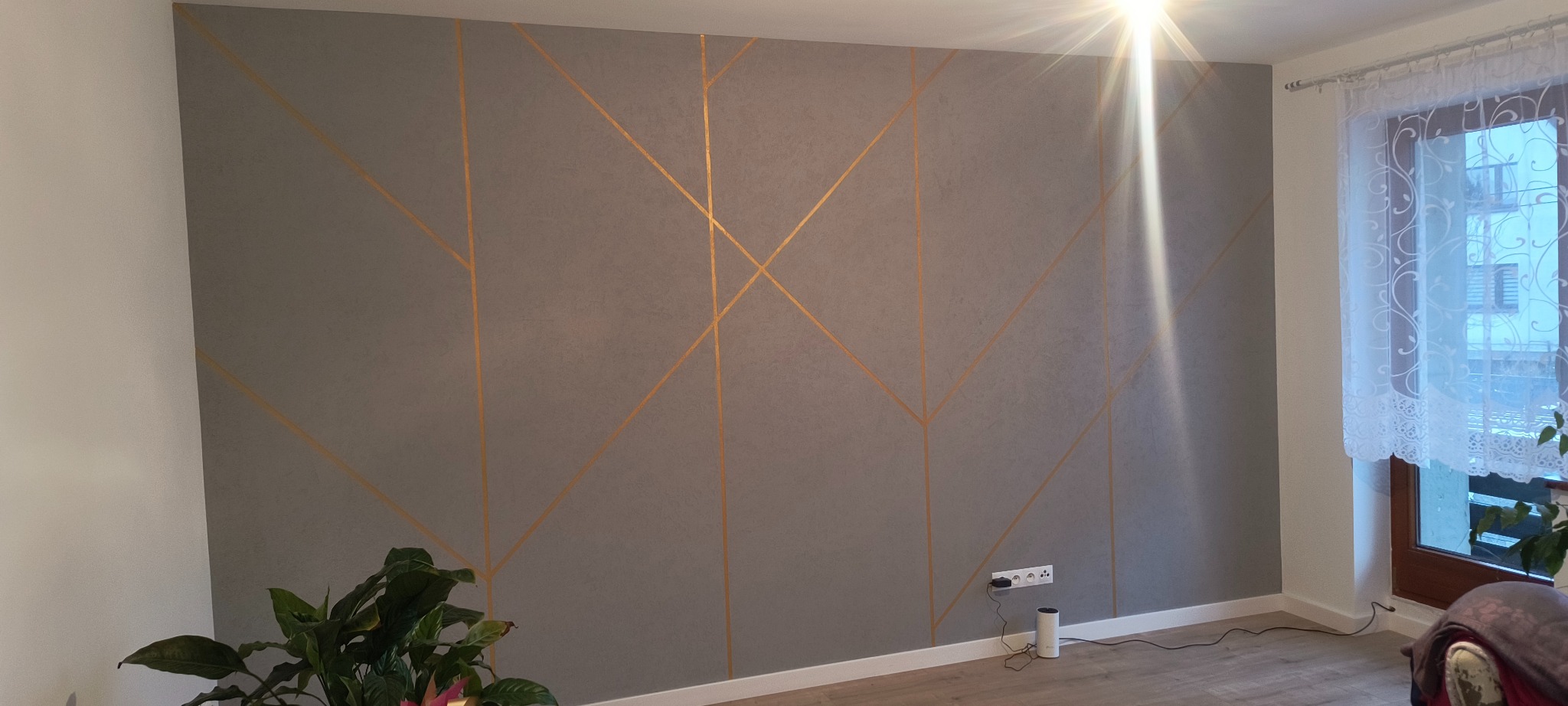 Szara ściana z geometrycznym wzorem z złotych linii, gniazdko elektryczne, roślina doniczkowa, okno z firanką i fragment podłogi.