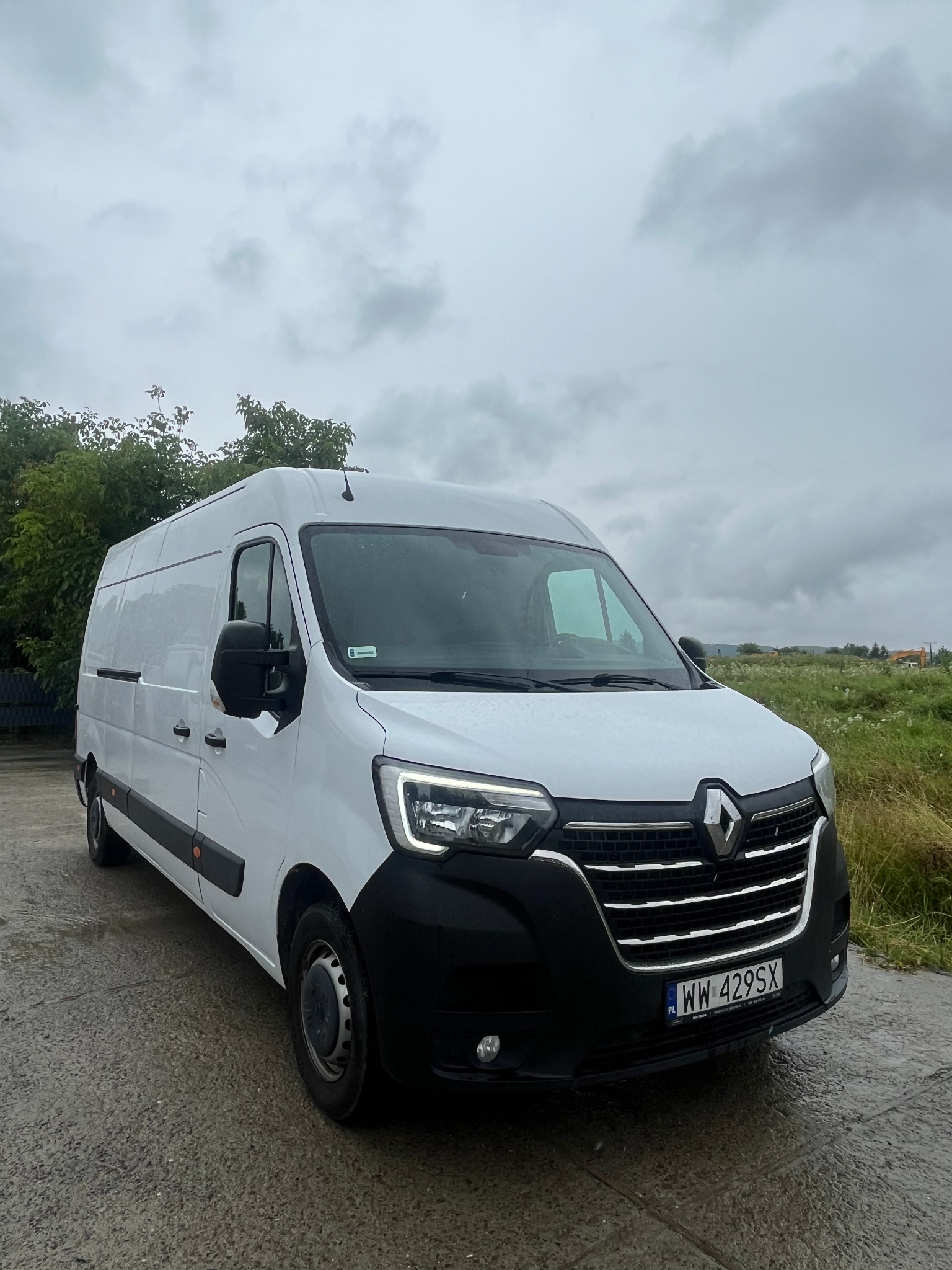 Biały Renault Master na mokrym asfalcie, tablica rejestracyjna WW 429SX, pochmurne niebo, widok z boku, zieleń w tle, transport.