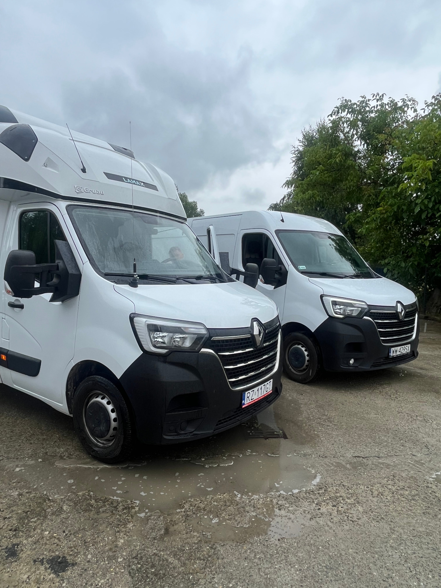 Dwa białe busy Renault Master z zabudową LAMAX, zaparkowane na mokrym placu w pochmurny dzień. Widoczne tablice rejestracyjne. Jeden bus z anteną.