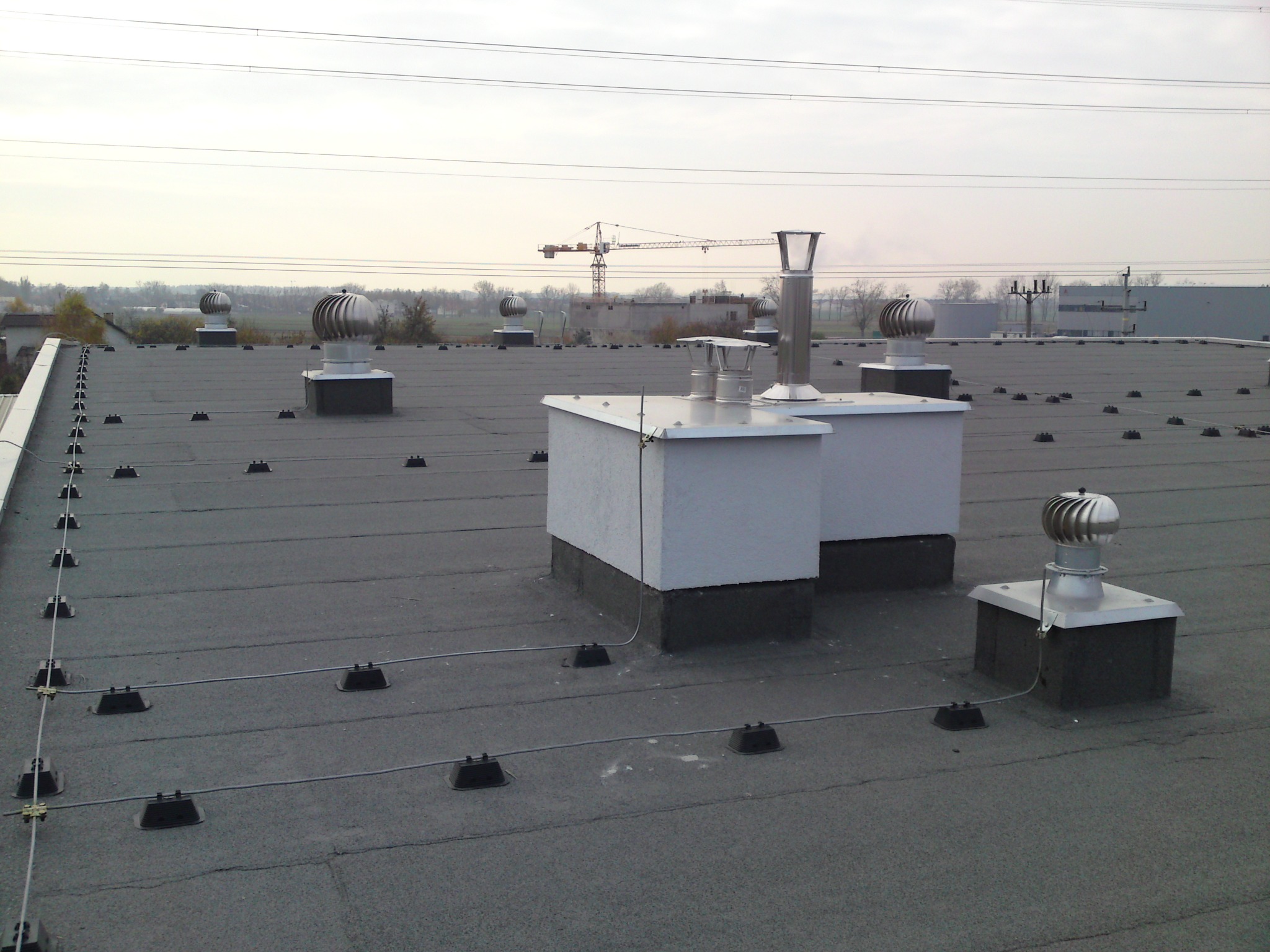 Szary dach przemysłowy z wentylatorami turbinowymi i elementami instalacji odgromowej, w tle widoczny żuraw budowlany.