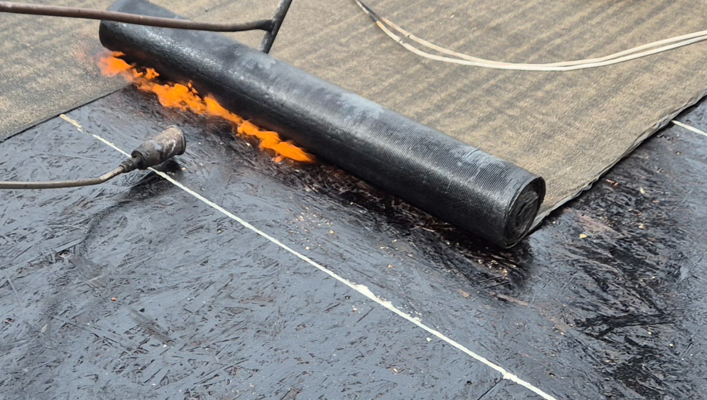 Prace dekarskie: palnik w akcji, zgrzewanie papy termozgrzewalnej na dachu pokrytym deskami. Widoczne rolka papy i płomienie.