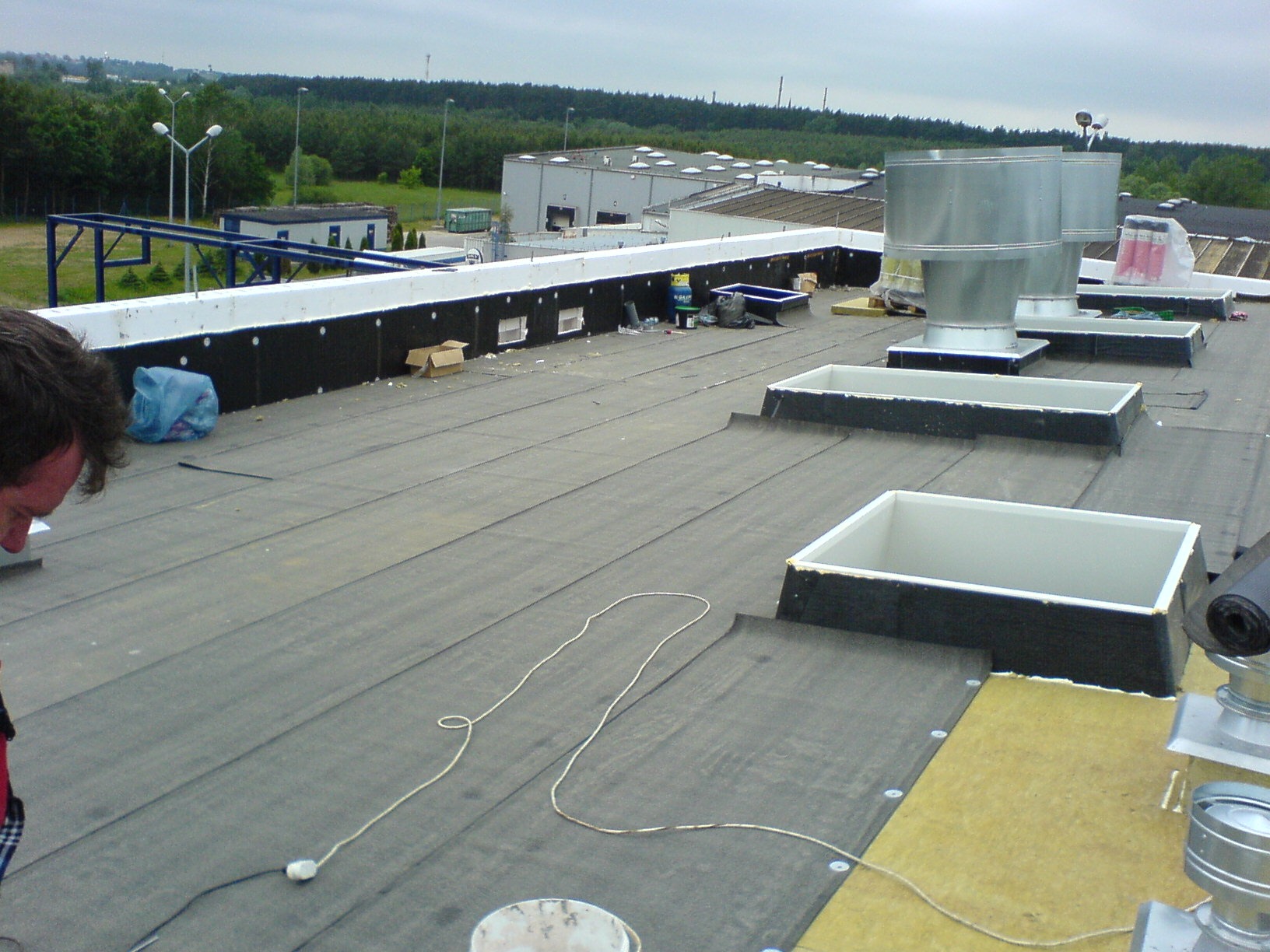 Rozległy dach płaski z papy termozgrzewalnej w trakcie prac. Widoczne świetliki dachowe, kominy wentylacyjne i częściowo ułożona izolacja. W tle budynek przemysłowy.