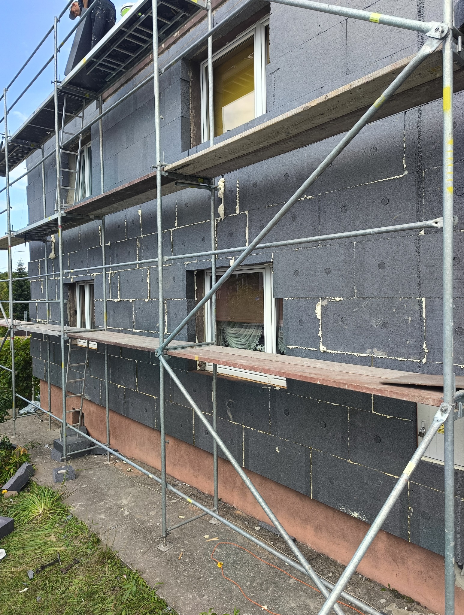 Dom w trakcie ocieplania: czarny styropian na elewacji, widoczne rusztowania. Prace w toku, pianka uszczelniająca między płytami.