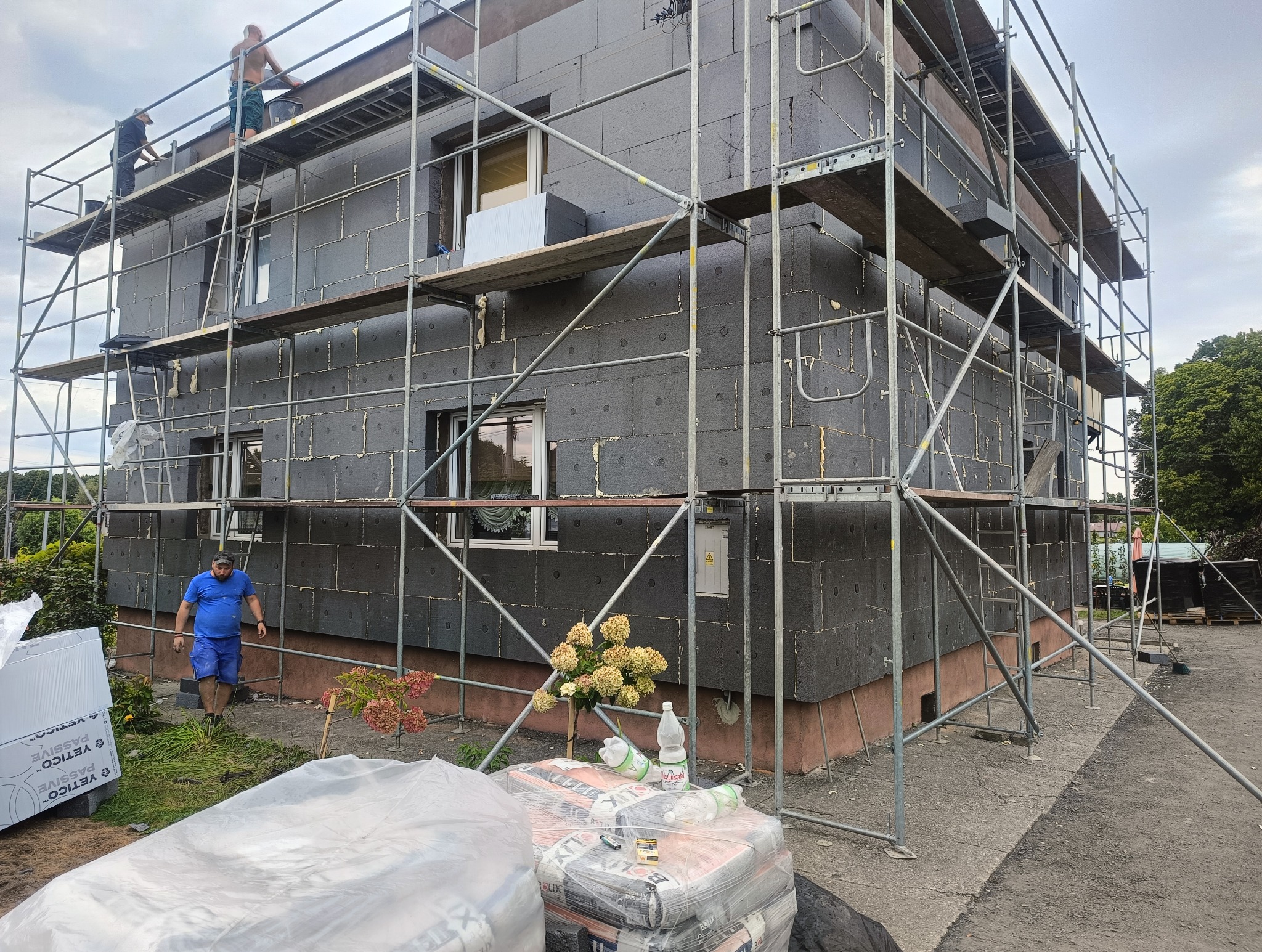 Dom w trakcie ocieplania, styropian w kolorze grafitowym na elewacji, rusztowania. Widoczni pracownicy na rusztowaniu i materiały budowlane.