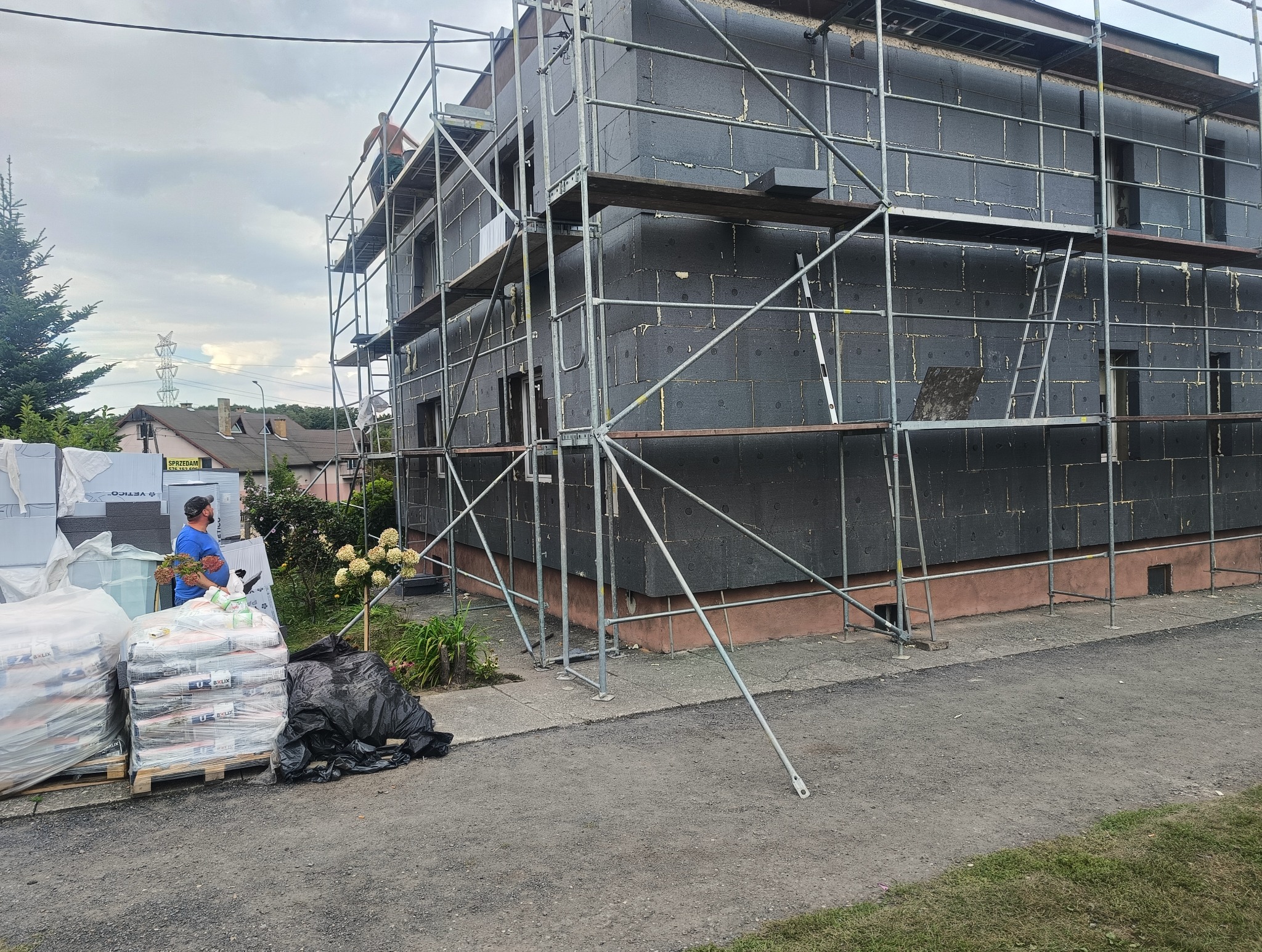 Dom w trakcie ocieplania elewacji, widoczne rusztowania i płyty izolacyjne, na dole paleta z workami materiałów budowlanych, oraz osoba w niebieskiej koszulce.