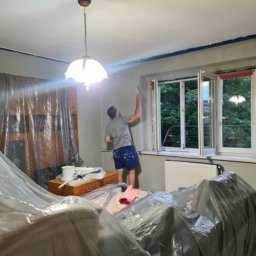 Rydzu - Malowanie ścian w pokoju z zabezpieczonymi meblami folią. Widoczny pracownik z wałkiem malarskim przy oknie i wiadro z farbą na komodzie.