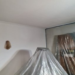 Rydzu - Pokój przygotowany do malowania: ściany z taśmą malarską, meble zakryte folią ochronną. Widoczny fragment sufitu i lampa na ścianie.
