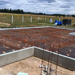 Fundament domu w trakcie budowy: zbrojenie, izolacja termiczna, elementy instalacji. Widok z góry na przygotowany szkielet konstrukcji na tle krajobrazu.